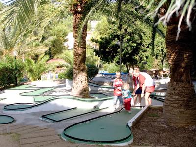 Eine Familie spielt Minigolf auf einem sonnigen, grünen Platz mit Palmen und Schatten.