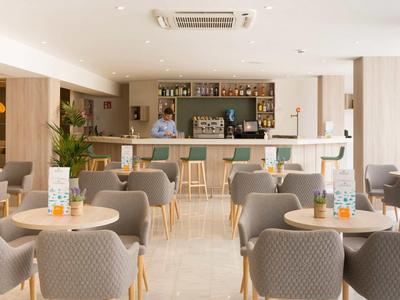 Helles Café mit grauen Stühlen, runden Tischen und einer Bar im Hintergrund mit einem Barkeeper.