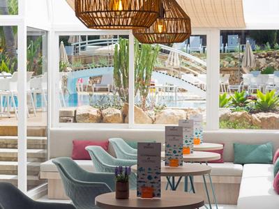 Helle Lounge mit pastellfarbenen Sesseln, runden Tischen und hängenden Holzlampen am Fenster.