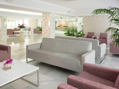 Helles Wohnzimmer mit grauem Sofa, mehreren rosa Sesseln, weißen Tischen und grünen Pflanzen.