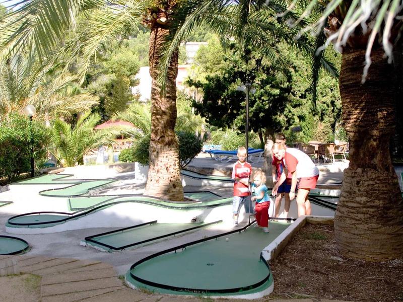 Famille jouant au mini-golf dans un jardin tropical ensoleillé avec des palmiers et des parcours verts.