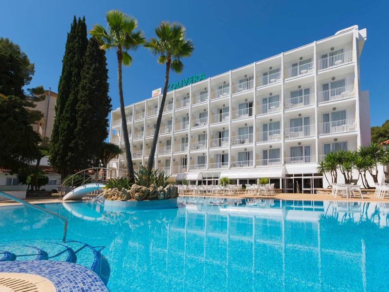 Hôtel moderne avec rangées de balcons donnant sur une grande piscine bleue entourée de palmiers.