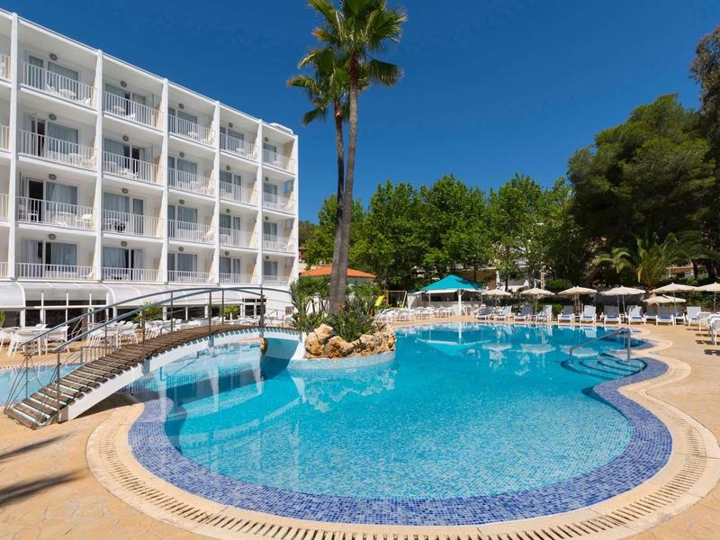 Piscine extérieure d'hôtel avec eau bleue claire, entourée de chaises longues et d'un bâtiment blanc.