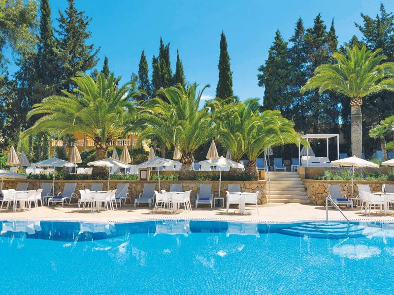 Zone de piscine avec transats, parasols et palmiers sous un ciel bleu ensoleillé.