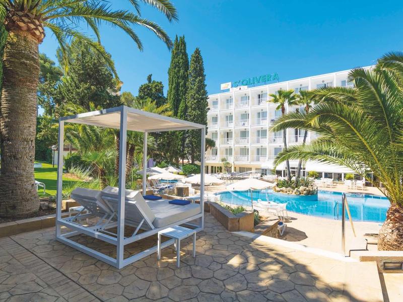 Espace piscine d'hôtel avec transats, palmiers et bâtiment blanc sous un ciel bleu clair.