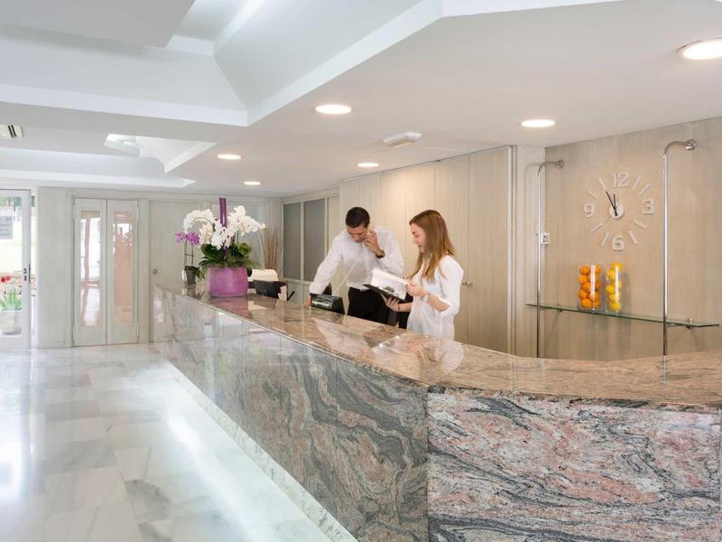 Réception d'hôtel avec comptoir en marbre, deux employés assistent une cliente, intérieur lumineux et moderne.