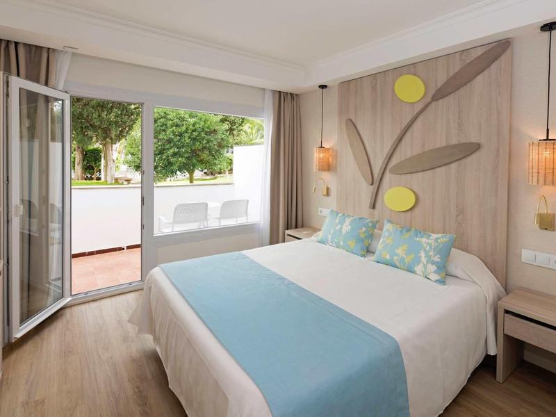 Chambre d'hôtel lumineuse avec lit double, accents bleus et porte ouverte sur terrasse verdoyante.