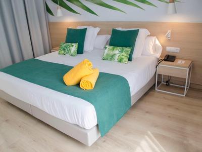 Nowoczesny pokój hotelowy z białym łóżkiem, turkusowym kocem, żółtymi ręcznikami i drewnianą podłogą.