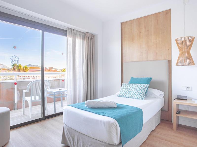 Camera d'hotel luminosa con letto singolo, biancheria blu e bianca e balcone con vista.