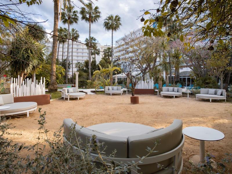 Area esterna moderna con panche rotonde e tavoli circondata da palme e edifici in vetro.