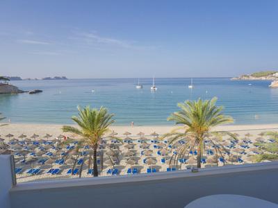 Beach with umbrellas, palm trees, and sailboats on calm sea under clear sky.