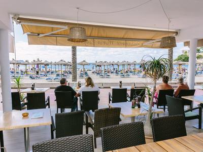 Covered beach bar with tables, chairs, and a view of the sandy beach and sea.