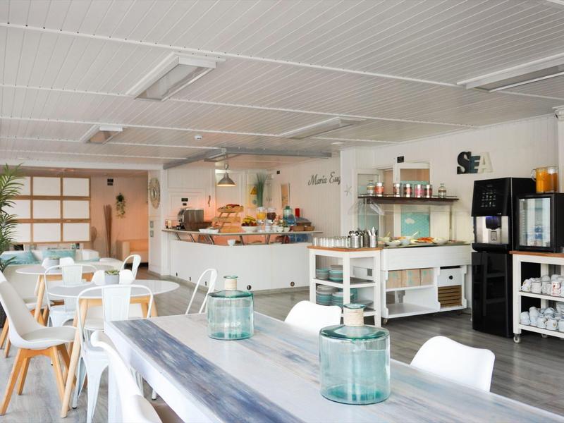 Bright dining area with white chairs, wooden tables, and a breakfast counter in the background.