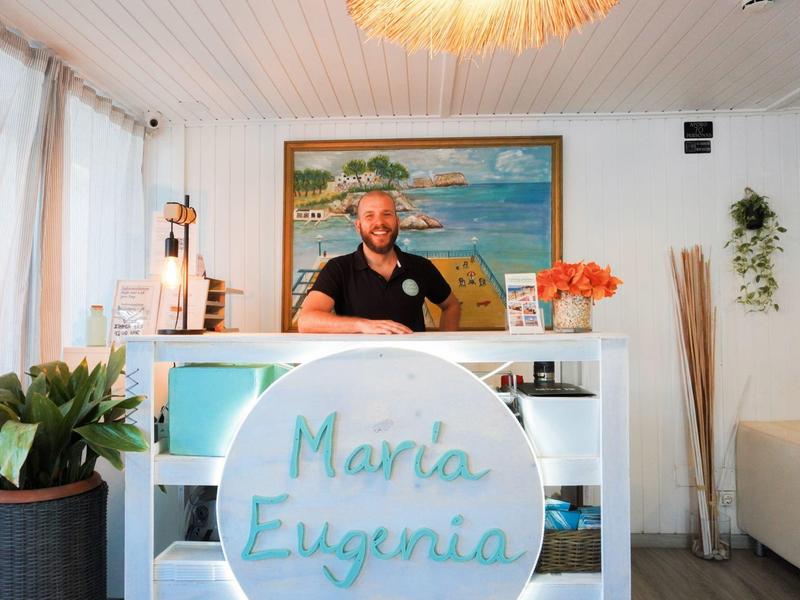 Hotel reception area with receptionist, plants, and beach painting on the wall.