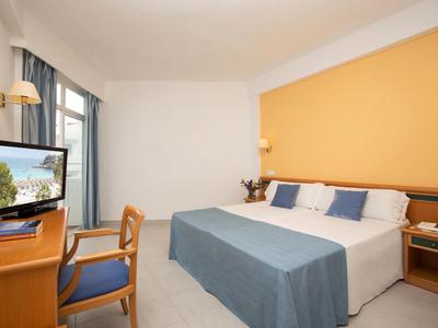 Schlichtes Hotelzimmer mit Doppelbett, blauem Bettüberwurf, Holzmöbeln und Fenster mit grauen Vorhängen.