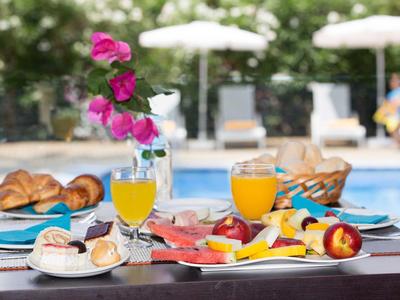 Tisch mit Frühstück, Croissants, Obst, Orangensaft und Blumen vor Pool mit Liegestühlen.