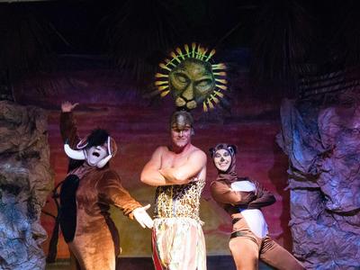 Drei Schauspieler in Tierkostümen und Sonnenmaske auf einer Bühne mit Felskulisse beim Theaterstück.