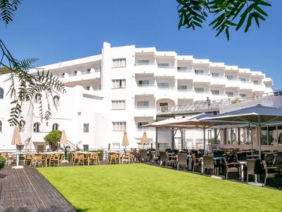 Modern hotel with terrace, seating areas, and green lawn under clear sky.