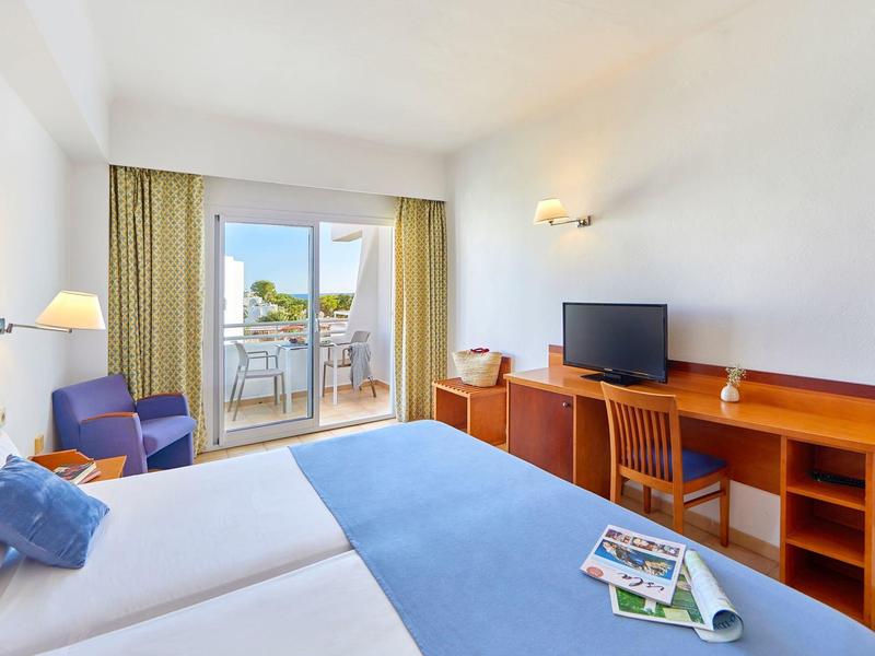Bright hotel room with twin beds, wooden furniture, TV, and balcony with outdoor seating.