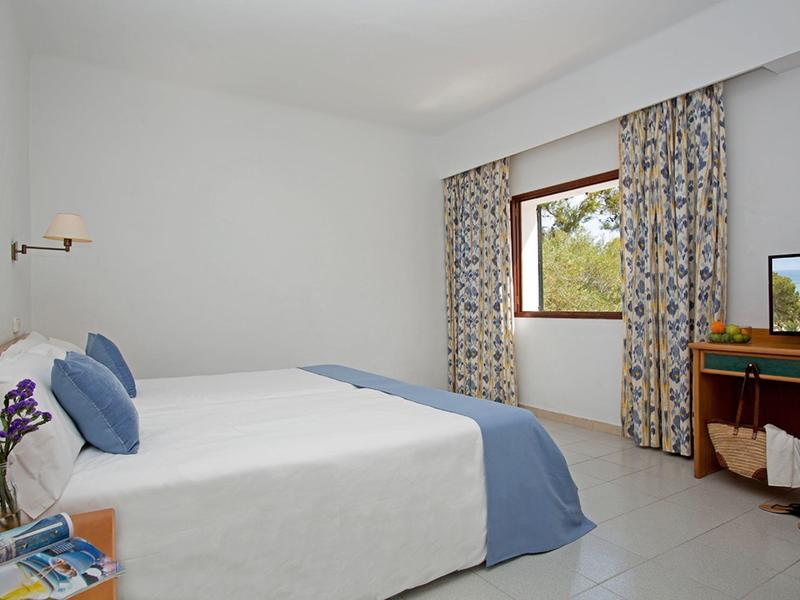 Bright hotel room with double bed, blue accents, window with floral curtains, and wooden desk.