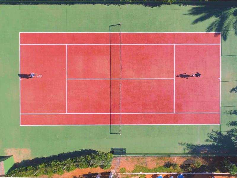 Red tennis court with players, surrounded by green grass and trees.