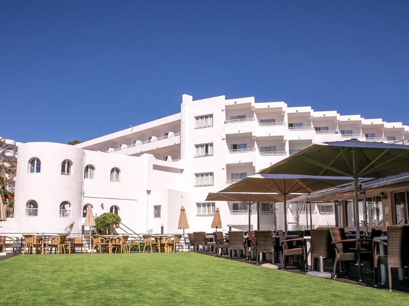 White hotel building with balconies beside lawn and outdoor seating with umbrellas.