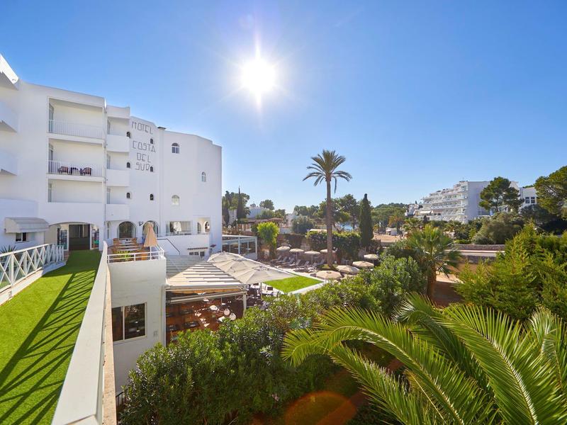 Sunny hotel garden with palm trees and white building under clear blue sky.
