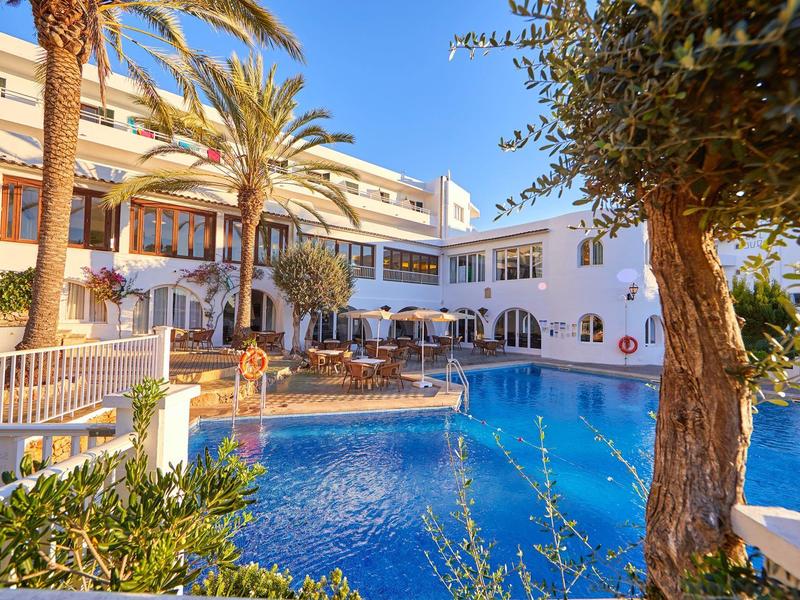Hotel with large pool, palm trees, and sun terrace under clear sky.