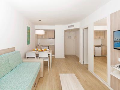 Habitación de hotel luminosa con sofá verde, mesa de comedor, cocina pequeña y espejo grande.