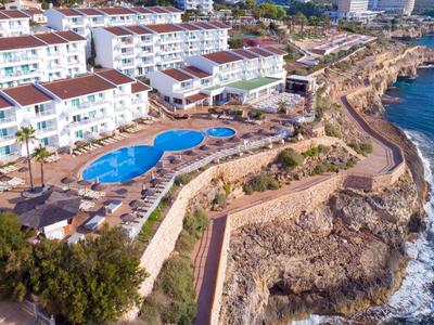 Complejo hotelero con piscina en la costa rocosa y vistas al mar azul bajo el sol.