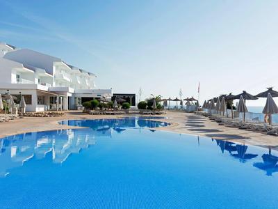 Piscina clara frente a edificio hotelero blanco con sombrillas y vista al mar en el horizonte.