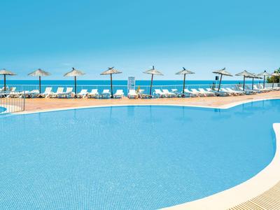 Piscine claire avec chaises longues et parasols face à la mer sous un ciel bleu clair.