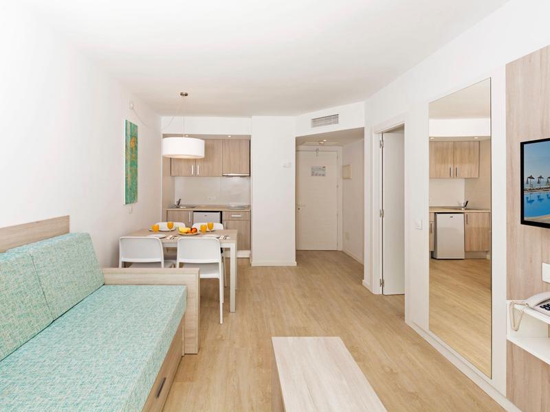 Habitación de hotel luminosa con sofá verde, mesa de comedor, cocina pequeña y espejo grande.