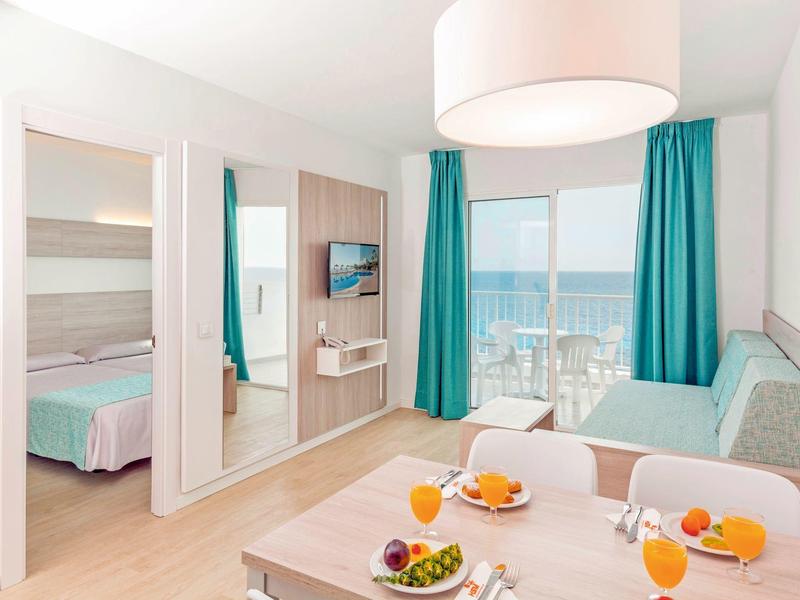 Habitación de hotel luminosa con mesa, sofá, vista al mar y grandes ventanas con cortinas azules.