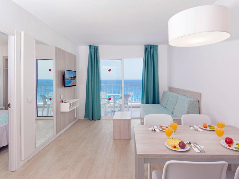 Habitación de hotel luminosa con vista al mar, mesa de comedor y asientos cómodos junto a la ventana.