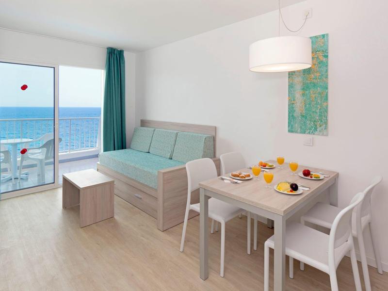 Sala de estar moderna con comedor, vista al mar y muebles claros de madera en una habitación de hotel.