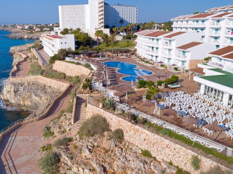 Complejo hotelero con piscinas sobre un acantilado frente al mar y un camino costero.