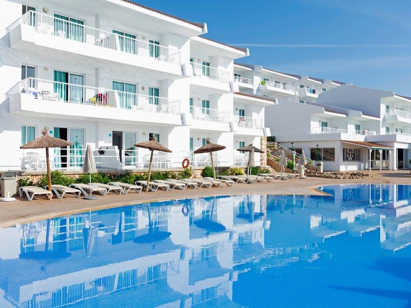 Complejo hotelero moderno de varios pisos con tumbonas y gran piscina en primer plano.