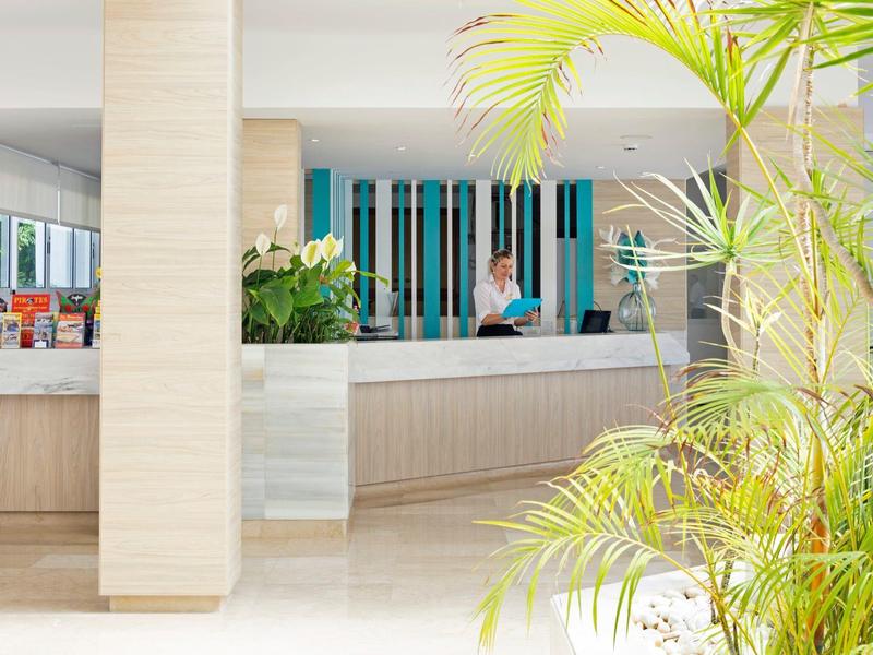 Recepción de hotel luminosa con plantas al frente y una persona atendiendo en el mostrador.