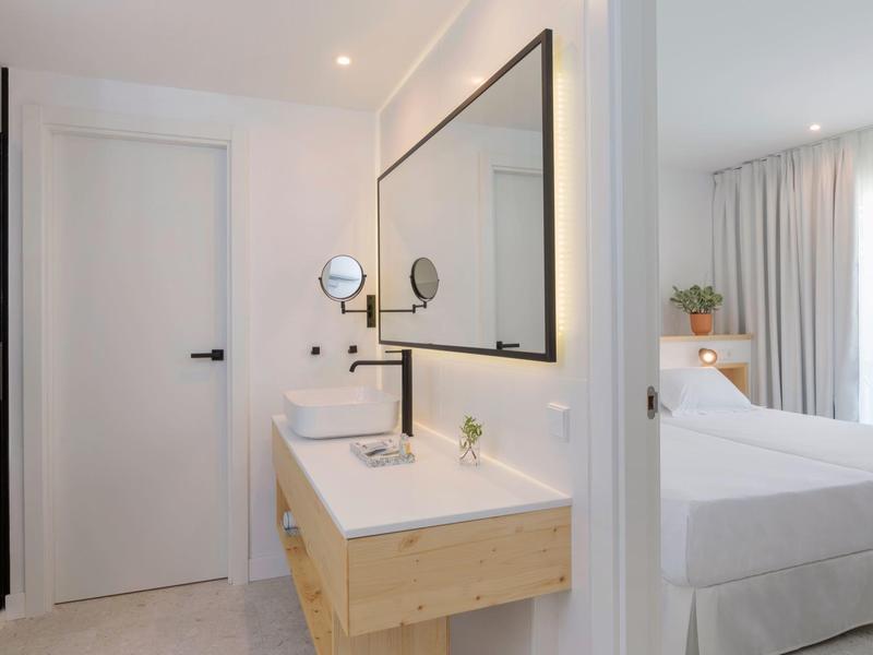 Bagno di hotel luminoso con doppi lavandini e grande specchio accanto a una camera da letto.