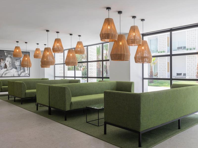 Area lounge moderna con divani verdi e lampade sospese accanto a grandi finestre.