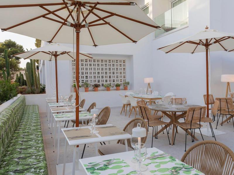 Area esterna moderna con tavoli bianchi, sedie in legno e grandi ombrelloni accanto a una fontana.