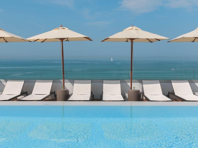 Vista della piscina con lettini e ombrelloni vicino al mare sotto un cielo azzurro.