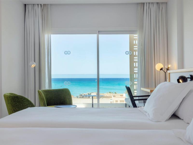 Camera d'hotel luminosa con letto bianco, sedia verde e vista sul mare attraverso ampia porta scorrevole in vetro.