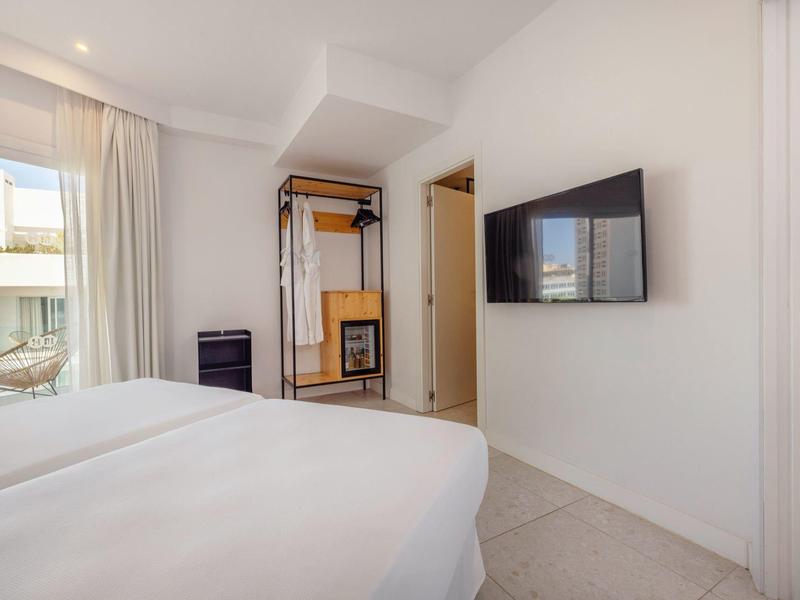 Camera d'albergo luminosa con due letti, TV a schermo piatto e guardaroba aperto accanto a una porta.