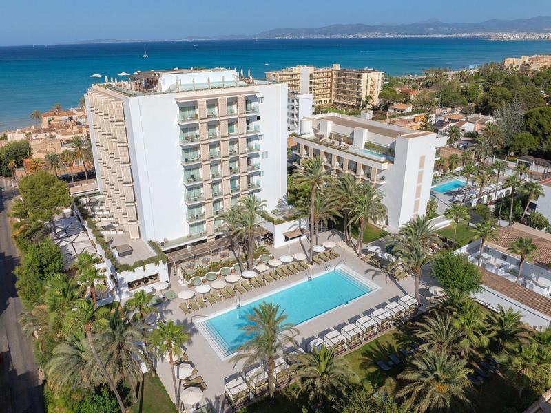 Hotel con piscina circondata da palme, vicino al mare e a una città sullo sfondo.