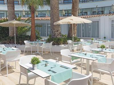 Area esterna luminosa di un hotel con tavoli e sedie bianchi sotto ombrelloni.