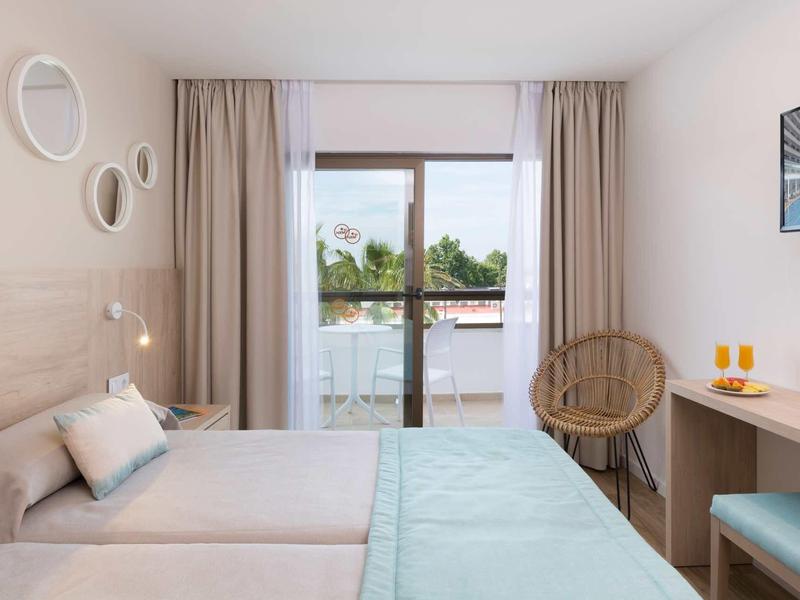 Habitación de hotel con dos camas, silla de mimbre, escritorio y balcón con vistas al lago.