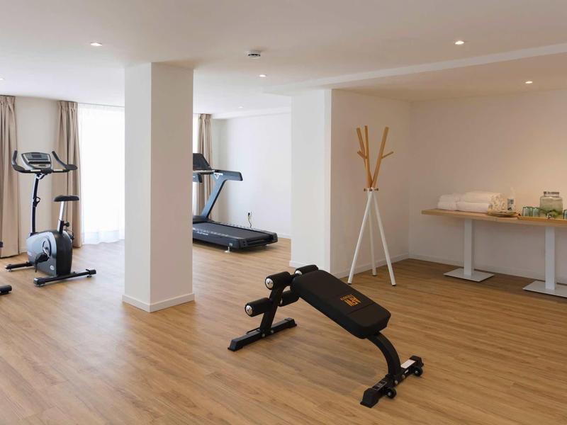 Gimnasio luminoso con cinta de correr, bicicleta, banco de entrenamiento y mesa de bebidas.