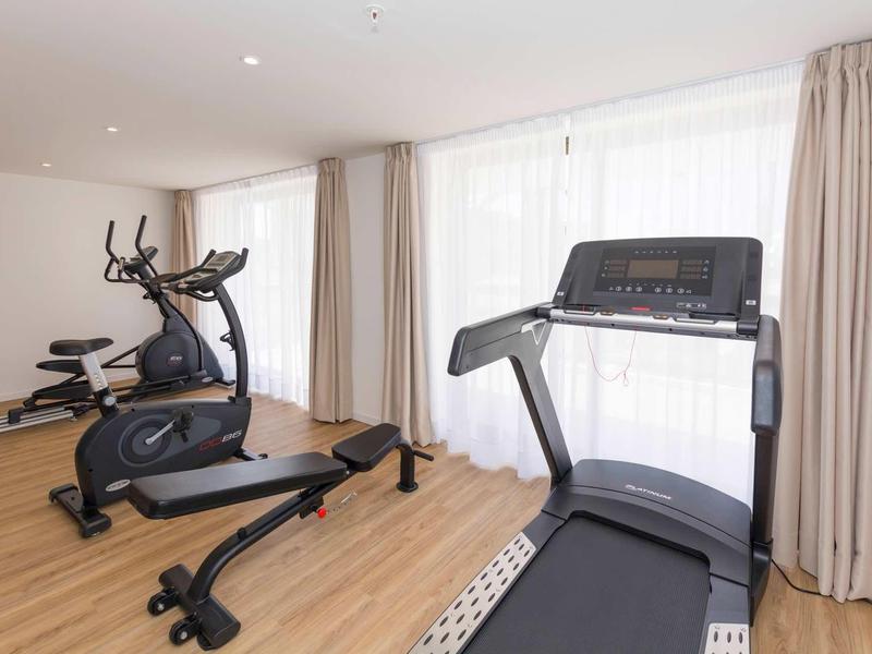 Gimnasio luminoso con cinta de correr, elíptica y banco de pesas frente a grandes ventanas con cortinas.